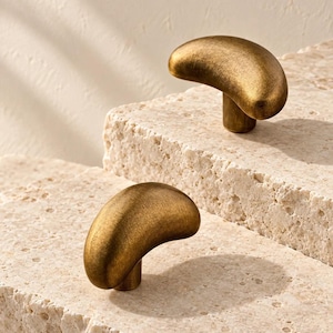 May include: Two brass cabinet knobs with a curved, bean-like shape and a brushed, matte finish. Each knob features a smooth, rounded top surface and a cylindrical base, showcasing a warm, metallic gold tone with subtle texture throughout.