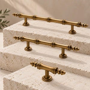 May include: Three brass-toned metal cabinet pulls with a bamboo-inspired design. The pulls feature a segmented cylindrical body with decorative finials at each end. The set includes one long pull with two posts and two shorter pulls of varying lengths.