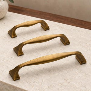 May include: Three arch-shaped cabinet pulls with a brushed antique brass finish. Each handle features a smooth, tapered center section that widens slightly, connecting to two angled, rectangular mounting bases on either end of the hardware.