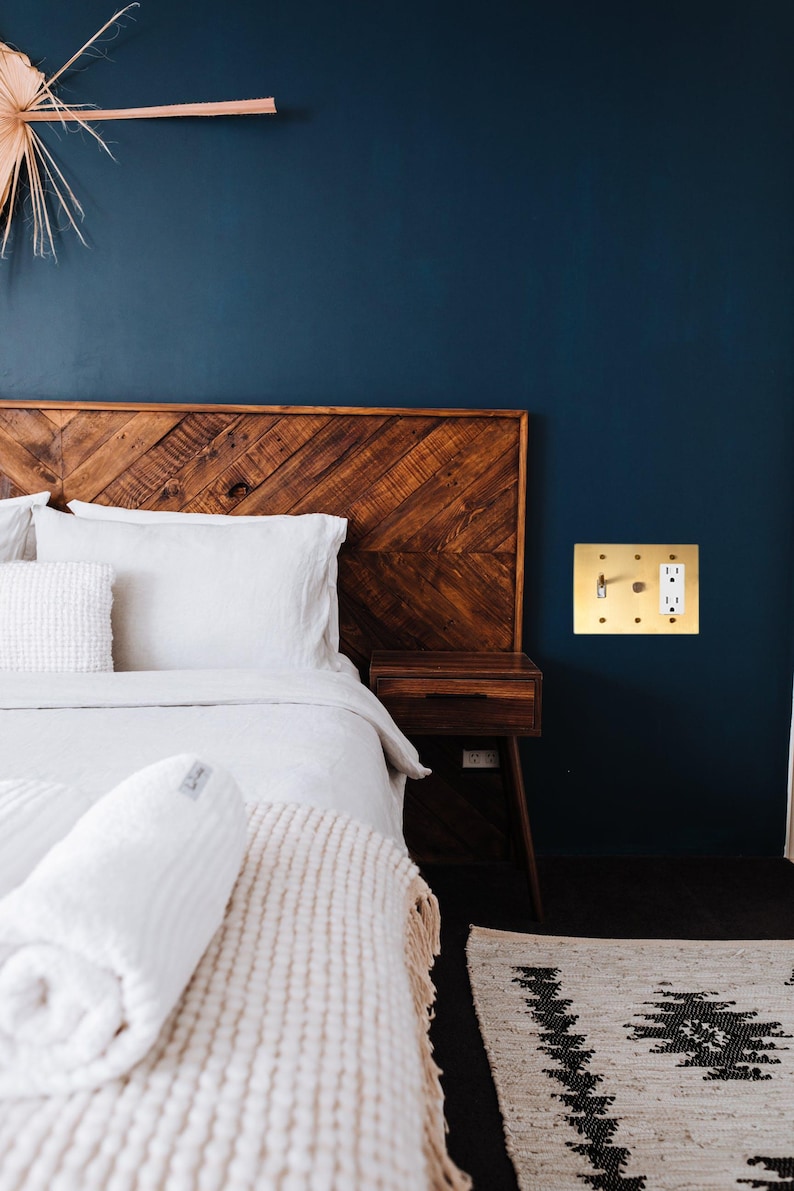 May include: A white bed with a wooden headboard and a white blanket. The bed is in a room with a dark blue wall and a gold light switch.