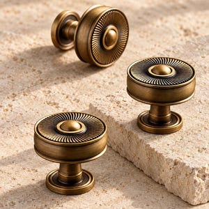 May include: Three round cabinet knobs made of antique brass with a brushed finish. Each knob features a central dome surrounded by a flat, circular face with fine, radiating grooves that extend from the center to the outer edge of the knob.