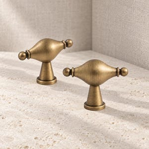 May include: Two brass-toned metal cabinet knobs featuring a central bulbous shape with small spherical ends and a tapered cylindrical base. The hardware has a brushed finish with visible metallic texture and a warm, muted golden color throughout.