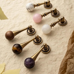 May include: A collection of six drop-style cabinet pulls featuring antique brass-toned metal hardware. Each pull is topped with a spherical accent in various materials, including white marble, pink quartz, clear quartz, dark wood, tiger eye, and amethyst.