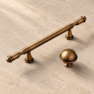 May include: A brushed antique brass cabinet handle with a long cylindrical bar and two mounting posts, accompanied by a matching round mushroom-shaped cabinet knob. Both hardware pieces feature a textured metallic finish and rounded ends.