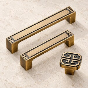 May include: Two rectangular cabinet pulls and one square cabinet knob in a brushed antique brass finish. Each piece features a dark, textured recessed border with a repeating geometric Greek key pattern at the corners and edges.