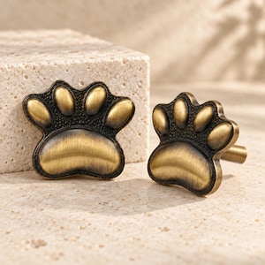 May include: Two paw print shaped cabinet knobs with a brushed antique brass finish. The knobs feature a textured dark background between the raised, smooth metallic toe pads and the large central pad, creating a distinct contrast.