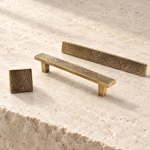 May include: A set of three brass-toned cabinet hardware pieces, including one square knob and two rectangular pulls. Each piece features a textured surface with a repeating pattern of etched, angled lines across the front and sides.
