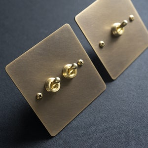 Puede incluir: Dos placas de interruptor de luz de latón con dos interruptores de palanca cada una. Las placas son cuadradas y tienen un acabado cepillado.