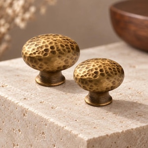 May include: Two oval-shaped cabinet knobs made of brass with a hammered, textured finish. Each knob features a rounded top surface with a dimpled pattern and a smooth, tapered base that connects to the mounting point, showcasing a metallic gold tone.