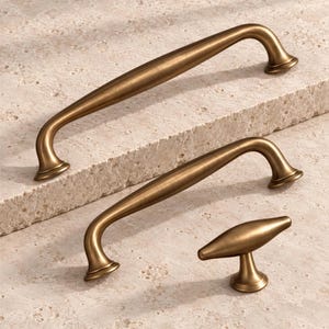 Pode incluir: Dois puxadores de armário alongados e curvos e um pequeno puxador de armário em forma de T, todos com acabamento em metal latão antigo escovado e liso. As ferragens apresentam um tom metálico quente e dourado com reflexos suaves e sutis.
