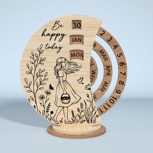 Puede incluir: Calendario perpetuo de madera con un dise&ntilde;o cortado con l&aacute;ser. El calendario presenta a una mujer caminando por un campo, las palabras "Be happy today" y anillos giratorios para la fecha, el mes y el d&iacute;a. El calendario es de madera clara.