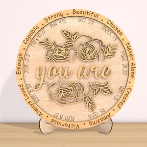 May include: A wooden round sign with the words "you are" in the center, surrounded by a floral design. The sign is decorated with bible verses and words of affirmation, such as "strong", "beautiful", "chosen", "capable", "enough", "always loved", "victorious", "amazing", "created", "never alone".