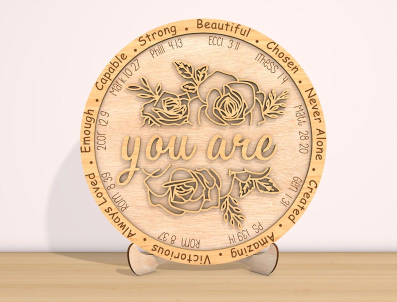 May include: A wooden round sign with the words "you are" in the center, surrounded by a floral design. The sign is decorated with bible verses and words of affirmation, such as "strong", "beautiful", "chosen", "capable", "enough", "always loved", "victorious", "amazing", "created", "never alone".