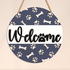 May include: Round wooden sign with a navy blue background featuring white paw prints and bone shapes. The word "Welcome" is in black script across a white stripe. A jute rope is attached for hanging.