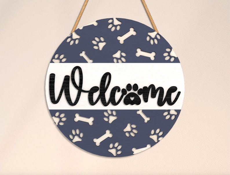 May include: Round wooden sign with a navy blue background featuring white paw prints and bone shapes. The word "Welcome" is in black script across a white stripe. A jute rope is attached for hanging.