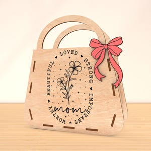 May include: Wooden cutout of a handbag with a pink bow. The handbag has the words "mom", "loved", "beautiful", "worthy", "important", and "strong" engraved on it.
