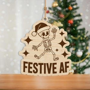 May include: Wooden Christmas ornament featuring a skeleton wearing a Santa hat, with the words "FESTIVE AF" in bold letters. The ornament has a light brown color and is hanging by a string. The background includes a Christmas tree.