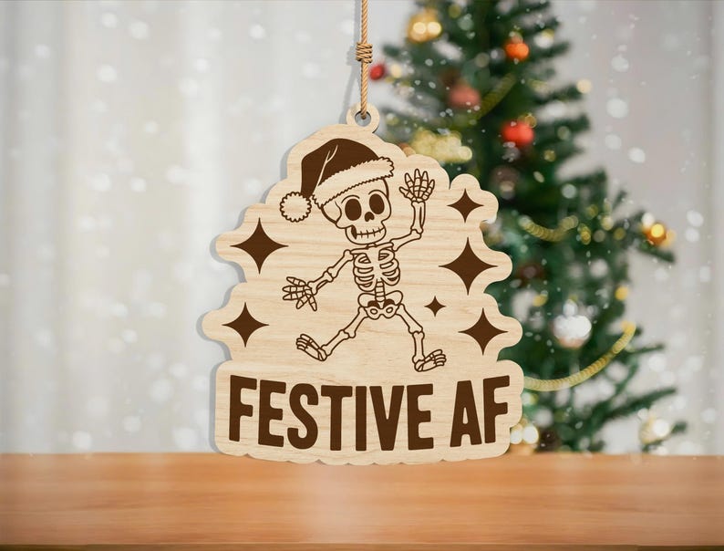 May include: Wooden Christmas ornament featuring a skeleton wearing a Santa hat, with the words "FESTIVE AF" in bold letters. The ornament has a light brown color and is hanging by a string. The background includes a Christmas tree.