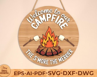 Könnte beinhalten: Runder Holzschild mit der Aufschrift "Welcome to our CAMPFIRE" und "THE S'MORE THE MERRIER". Es zeigt ein Lagerfeuer mit Flammen, Holzscheiten und Marshmallows an Stöcken. Das Schild hat eine Schnur zum Aufhängen.