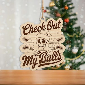 May include: Wooden Christmas ornament with a skeleton wearing a Santa hat and holding ornaments. The ornament has the text "Check Out My Balls" in a brown, vintage font. A rope is attached for hanging.