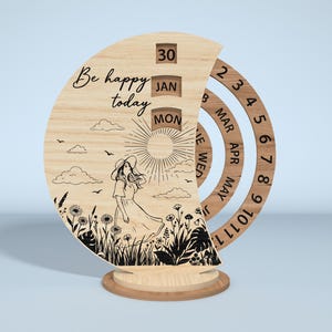 Puede incluir: Calendario perpetuo de madera con un dise&ntilde;o semicircular. El calendario presenta una ilustraci&oacute;n grabada con l&aacute;ser de una mujer en un campo, el texto "Be happy today" y anillos de fecha giratorios. La fecha muestra 30, JAN y MON.