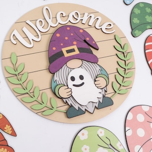 K&ouml;nnte beinhalten: Ein rundes Holzschild mit dem Wort Welcome in wei&szlig;er Schrift. In der Mitte ist ein Gnom mit violettem Hut, grauem Bart und gr&uuml;nen &Auml;rmeln abgebildet, der eine kleine wei&szlig;e Geisterfigur h&auml;lt. Zwei gr&uuml;ne Blattzweige rahmen den Gnom seitlich ein.