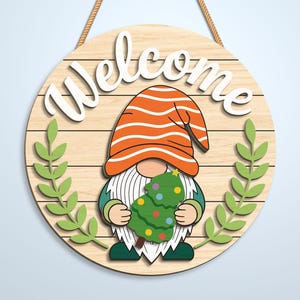 K&ouml;nnte beinhalten: Ein rundes Holzschild mit heller Holzmaserung und dem wei&szlig;en Schriftzug Welcome im oberen Bereich. Ein Gnom mit orangefarben gestreifter M&uuml;tze und langem wei&szlig;en Bart h&auml;lt einen gr&uuml;nen Weihnachtsbaum mit bunten Ornamenten, flankiert von zwei gr&uuml;nen Blattzweigen.