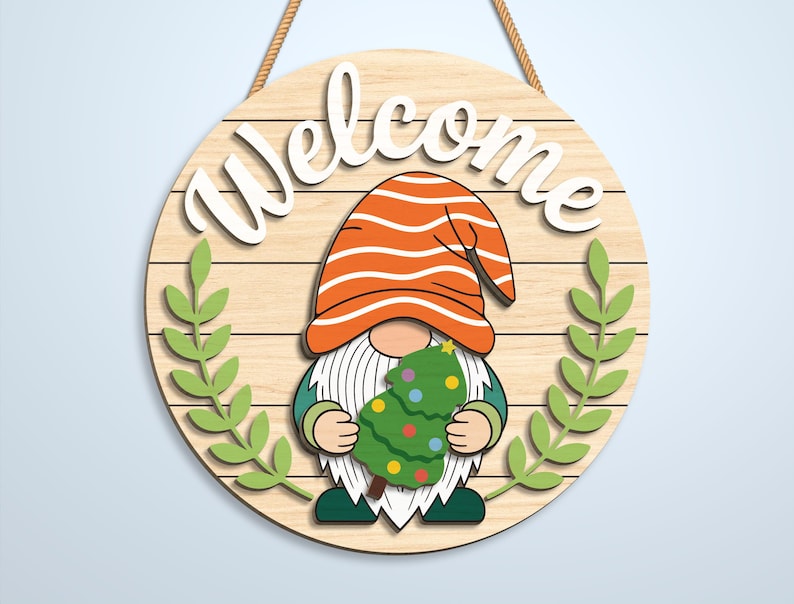 K&ouml;nnte beinhalten: Ein rundes Holzschild mit heller Holzmaserung und dem wei&szlig;en Schriftzug Welcome im oberen Bereich. Ein Gnom mit orangefarben gestreifter M&uuml;tze und langem wei&szlig;en Bart h&auml;lt einen gr&uuml;nen Weihnachtsbaum mit bunten Ornamenten, flankiert von zwei gr&uuml;nen Blattzweigen.