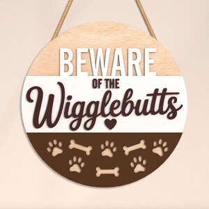May include: Round wooden sign with the text "BEWARE of the Wigglebutts". The sign is divided into three sections: light wood, white, and brown. The bottom section features paw prints and bone shapes.