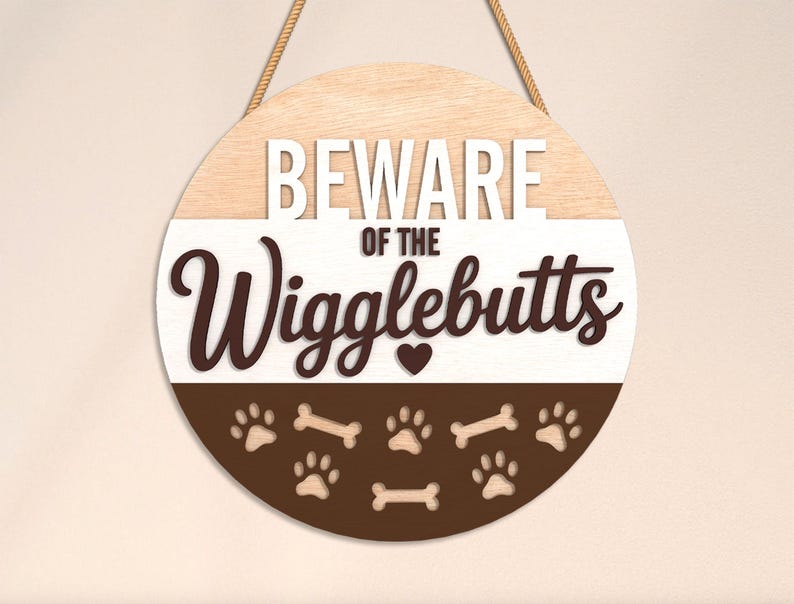 May include: Round wooden sign with the text "BEWARE of the Wigglebutts". The sign is divided into three sections: light wood, white, and brown. The bottom section features paw prints and bone shapes.