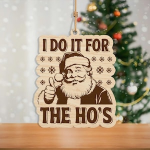 May include: Wooden Christmas ornament featuring Santa Claus with a thumbs-up gesture. The ornament has the text "I DO IT FOR THE HO'S" in a brown color. The ornament is hanging from a string.