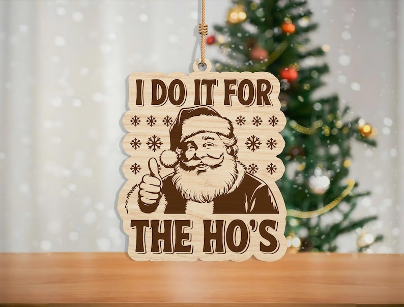 May include: Wooden Christmas ornament featuring Santa Claus with a thumbs-up gesture. The ornament has the text "I DO IT FOR THE HO'S" in a brown color. The ornament is hanging from a string.