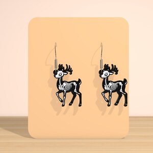 May include: A pair of black deer skeleton earrings with white bone details. The earrings are hanging on a peach-colored display card. The deer have antlers and are suspended from silver-colored hooks.