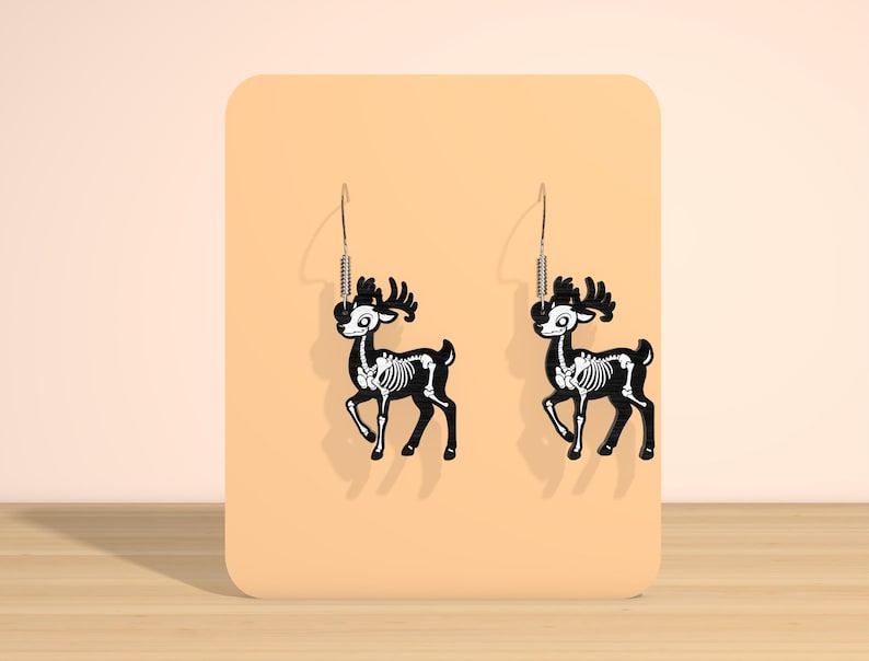 May include: A pair of black deer skeleton earrings with white bone details. The earrings are hanging on a peach-colored display card. The deer have antlers and are suspended from silver-colored hooks.