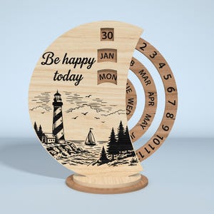 Puede incluir: Calendario perpetuo de madera con dise&ntilde;o de faro y velero. El calendario muestra la fecha, el mes y el d&iacute;a, con el texto "Be happy today". El calendario es de madera clara y tiene una base redonda.