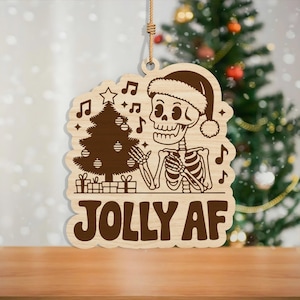 May include: Wooden Christmas ornament featuring a skeleton wearing a Santa hat, with a Christmas tree and musical notes. The text "JOLLY AF" is displayed in bold letters. The ornament is hanging by a natural twine.