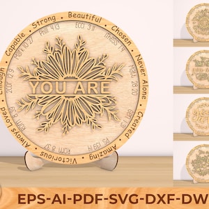 May include: A wooden round sign with a floral design and the words "YOU ARE" in the center. The sign is surrounded by a circle of words and bible verses, including "Strong", "Capable", "Beautiful", "Chosen", "Never Alone", "Created", "Amazing", "Victorious", "Always Loved", "Enough".