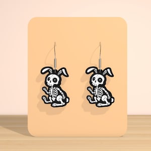 May include: A pair of black and white skeleton bunny earrings. The earrings feature a sitting bunny skeleton design with a black outline and white details. The earrings are displayed on a peach-colored background.