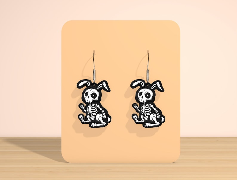 May include: A pair of black and white skeleton bunny earrings. The earrings feature a sitting bunny skeleton design with a black outline and white details. The earrings are displayed on a peach-colored background.