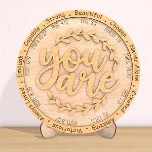 May include: A round wooden plaque with the words "You are" in a decorative font. The plaque is surrounded by a wreath of leaves and has various bible verses and words of affirmation around the edge, including "Strong", "Beautiful", "Chosen", "Never Alone", "Created", "Amazing", "Victorious", "Always Loved", "Enough", "Capable".