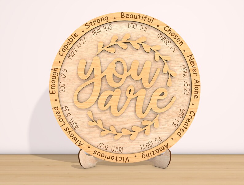 May include: A round wooden plaque with the words "You are" in a decorative font. The plaque is surrounded by a wreath of leaves and has various bible verses and words of affirmation around the edge, including "Strong", "Beautiful", "Chosen", "Never Alone", "Created", "Amazing", "Victorious", "Always Loved", "Enough", "Capable".