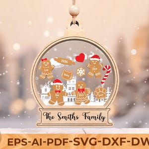 Op de afbeelding: Een houten kerstversiering in de vorm van een sneeuwbol, met peperkoekenmannetjes, een hart en een zuurstok. De versiering is voorzien van de tekst "The Smiths Family" en het jaar "2026".