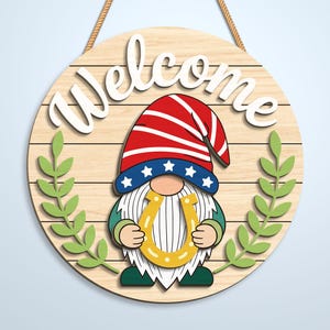 K&ouml;nnte beinhalten: Ein rundes Holzschild mit Kordelaufh&auml;ngung und dem Schriftzug Welcome in wei&szlig;er Schreibschrift. In der Mitte ist ein Wichtel mit rot-wei&szlig;-blauem Sternenhut abgebildet, der ein gelbes Hufeisen h&auml;lt. Zwei gr&uuml;ne Zweige rahmen den Wichtel ein.