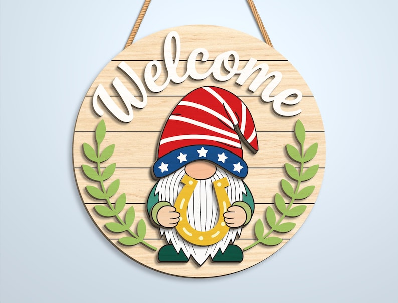 K&ouml;nnte beinhalten: Ein rundes Holzschild mit Kordelaufh&auml;ngung und dem Schriftzug Welcome in wei&szlig;er Schreibschrift. In der Mitte ist ein Wichtel mit rot-wei&szlig;-blauem Sternenhut abgebildet, der ein gelbes Hufeisen h&auml;lt. Zwei gr&uuml;ne Zweige rahmen den Wichtel ein.