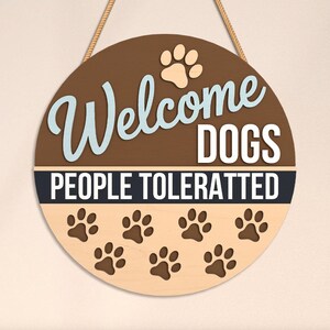 May include: Round wooden sign with the text "Welcome DOGS PEOPLE TOLERATTED". The sign has a brown background with light blue and white lettering, paw prints, and a rope for hanging.