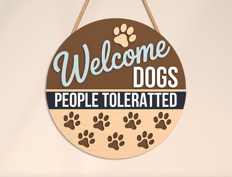 May include: Round wooden sign with the text "Welcome DOGS PEOPLE TOLERATTED". The sign has a brown background with light blue and white lettering, paw prints, and a rope for hanging.