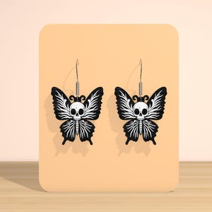 May include: A pair of black butterfly earrings with a white skull design. The butterfly wings have a detailed, stylized pattern. The earrings hang from silver-colored hooks.