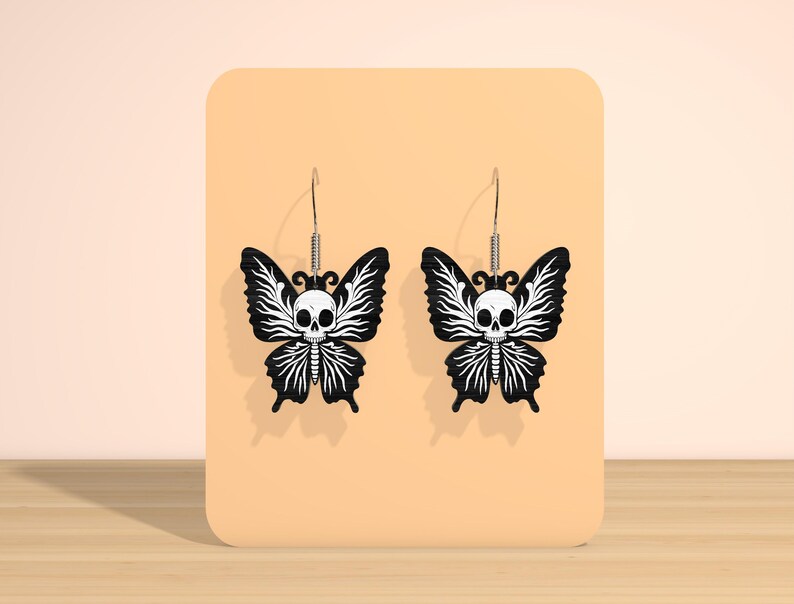 May include: A pair of black butterfly earrings with a white skull design. The butterfly wings have a detailed, stylized pattern. The earrings hang from silver-colored hooks.