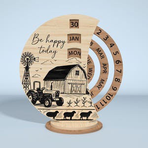 Puede incluir: Calendario perpetuo de madera con una escena de granja, que incluye un tractor, un granero y ovejas. El calendario muestra la fecha, el mes y el d&iacute;a, con las palabras "Be happy today" grabadas en la parte delantera. La base es redonda.