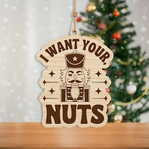 May include: Wooden Christmas ornament with the text "I WANT YOUR, NUTS" and a nutcracker design. The ornament is brown and hangs from a natural twine string. A Christmas tree is visible in the background.